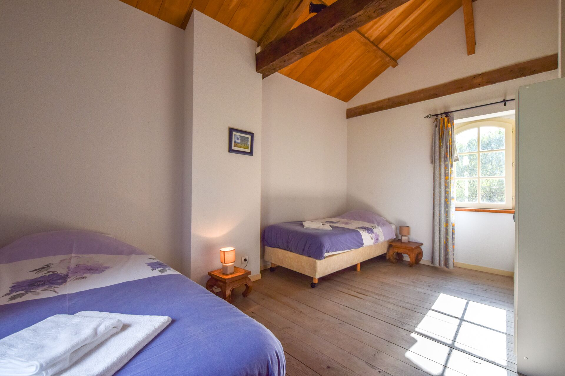 A bedroom with two beds, wooden flooring, a window with curtains, and a small bedside table with a lamp.