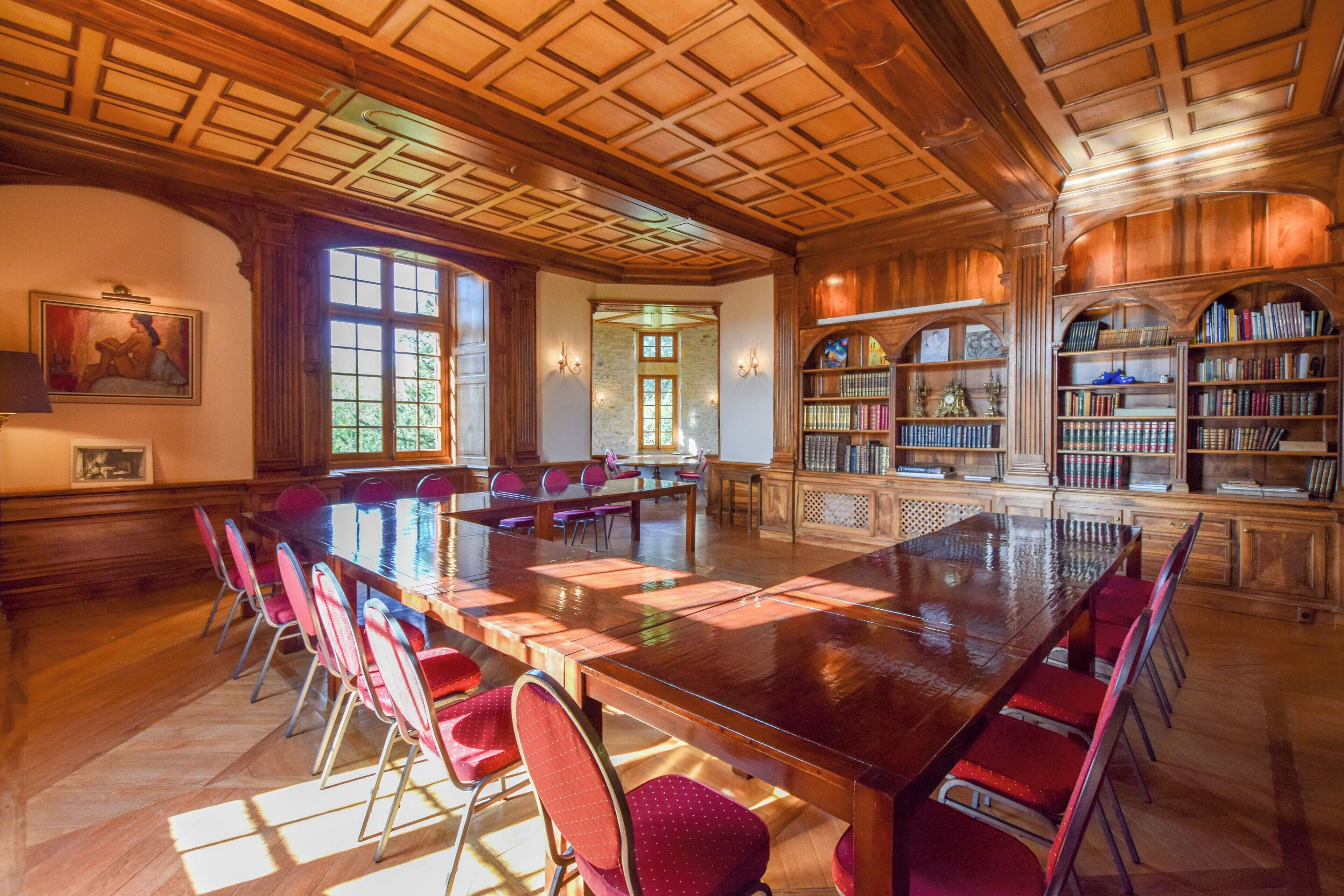 Une bibliothèque spacieuse, avec une grande table en bois et des chaises rouges, entourée d’étagères.