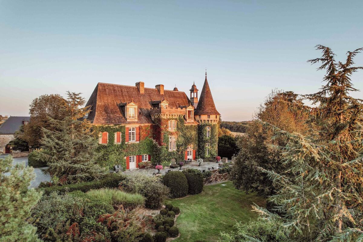 Un edificio grande e histórico con múltiples torres y muros cubiertos de hiedra, rodeado de jardines y árboles bien cuidados.