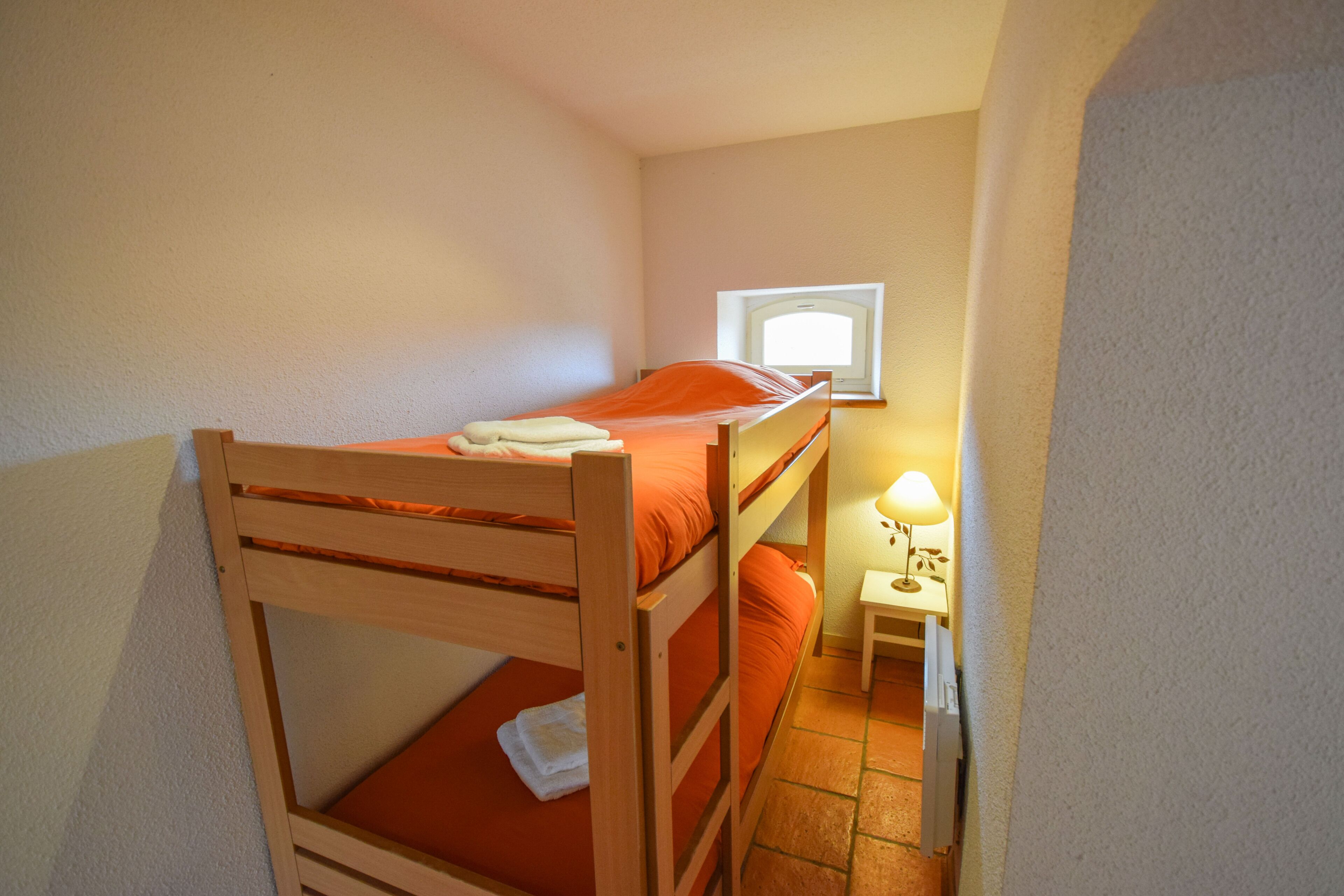 A small, single-bedded room with a wooden bunk bed, a bedside table with a lamp, and a window providing natural light.