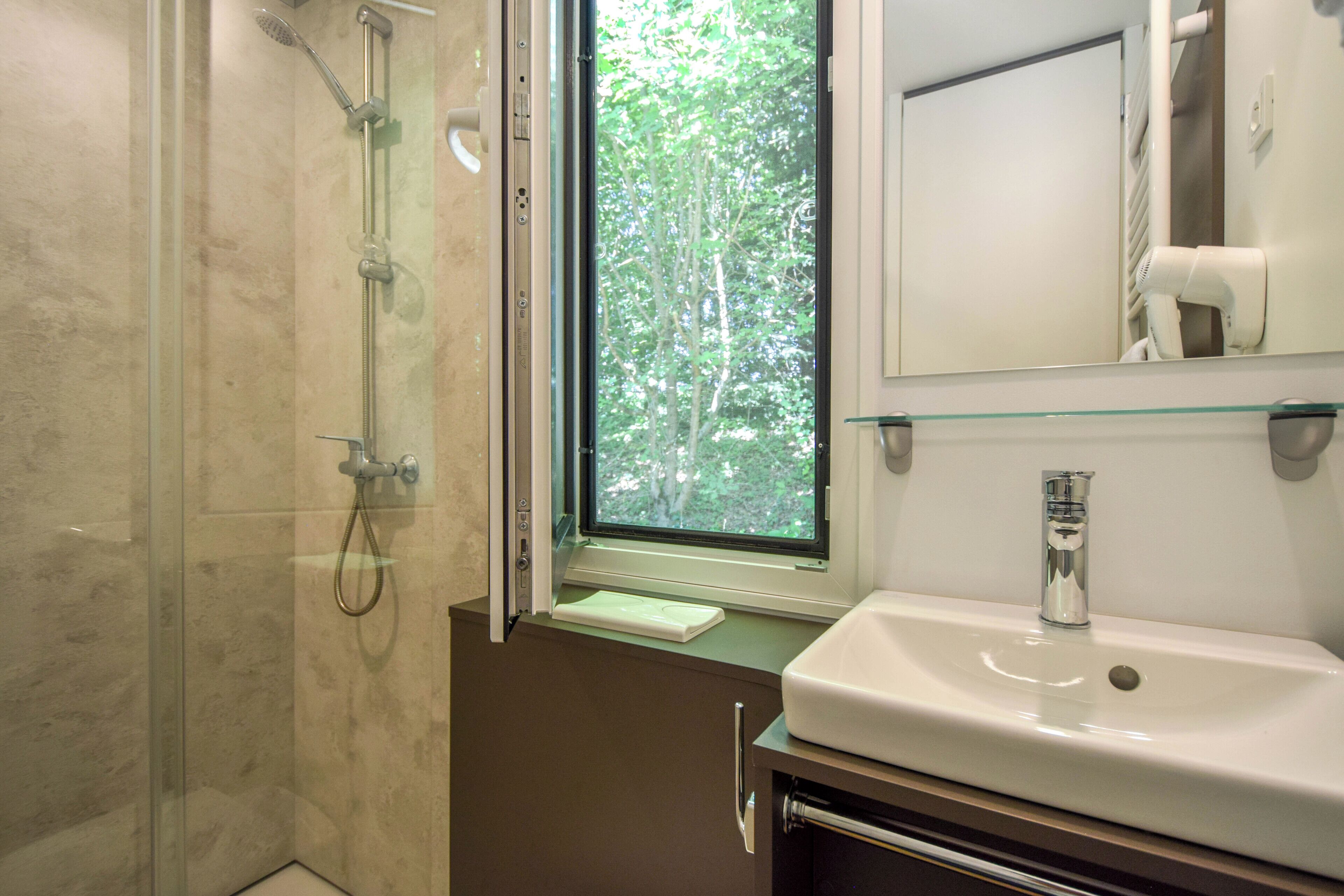 A bathroom with a glass-enclosed shower, a window with a view of trees, a white sink, and a mirror above the sink.