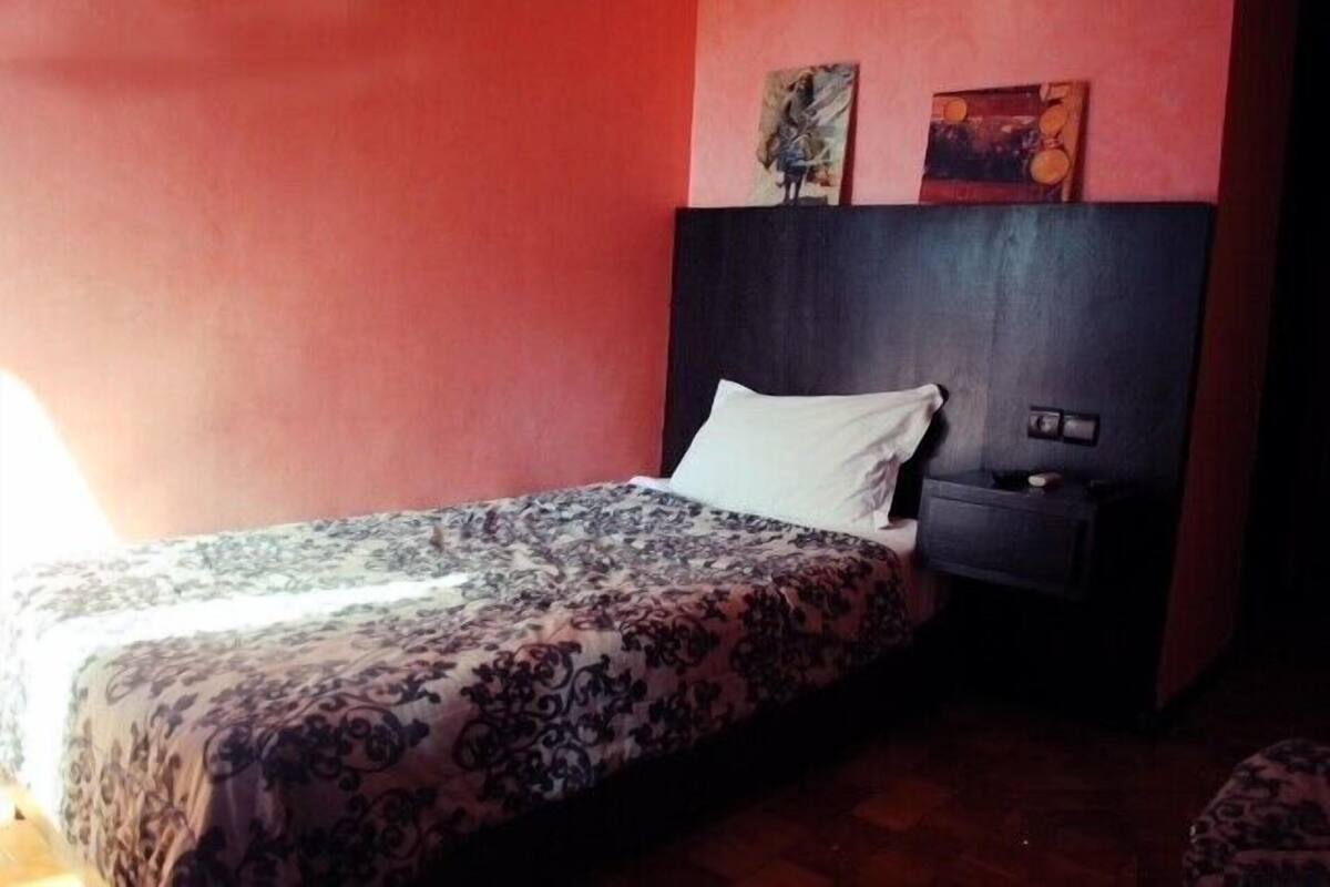 Una cama individual con una colcha estampada, una mesita de noche con lámpara, y dos cuadros en la pared sobre la cama.