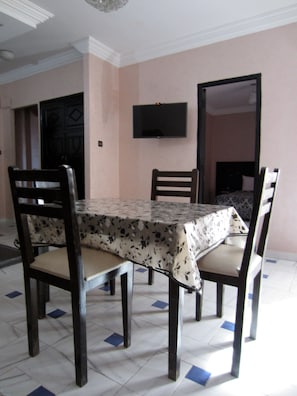 Un comedor con una mesa cubierta por un mantel floral, rodeada de sillas de madera con asientos acolchados. Un televisor de pantalla plana está montado en la pared sobre la mesa.