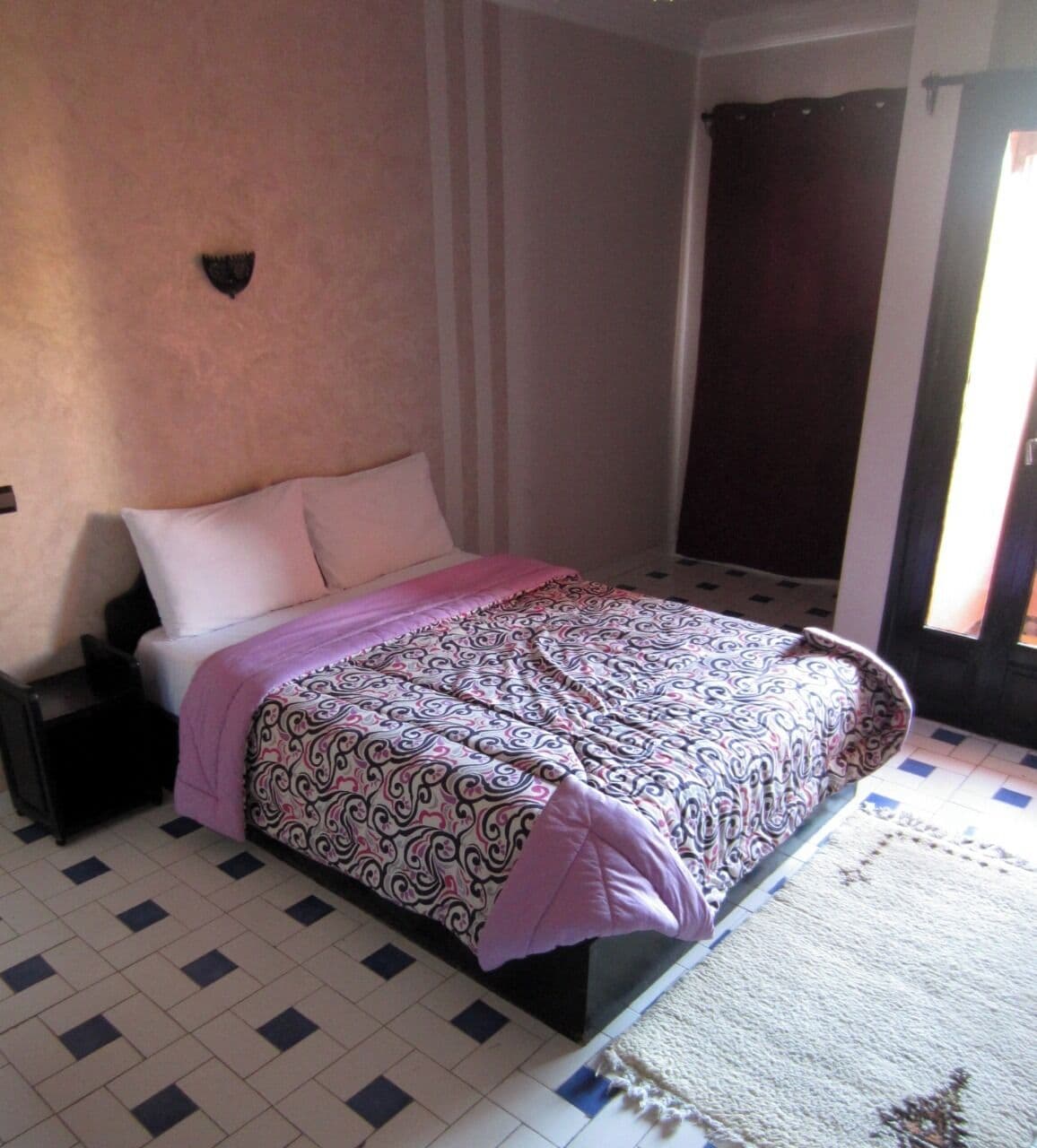 Un dormitorio con cama, mesita de noche, lámpara de pared, ventana con cortinas y alfombra estampada.