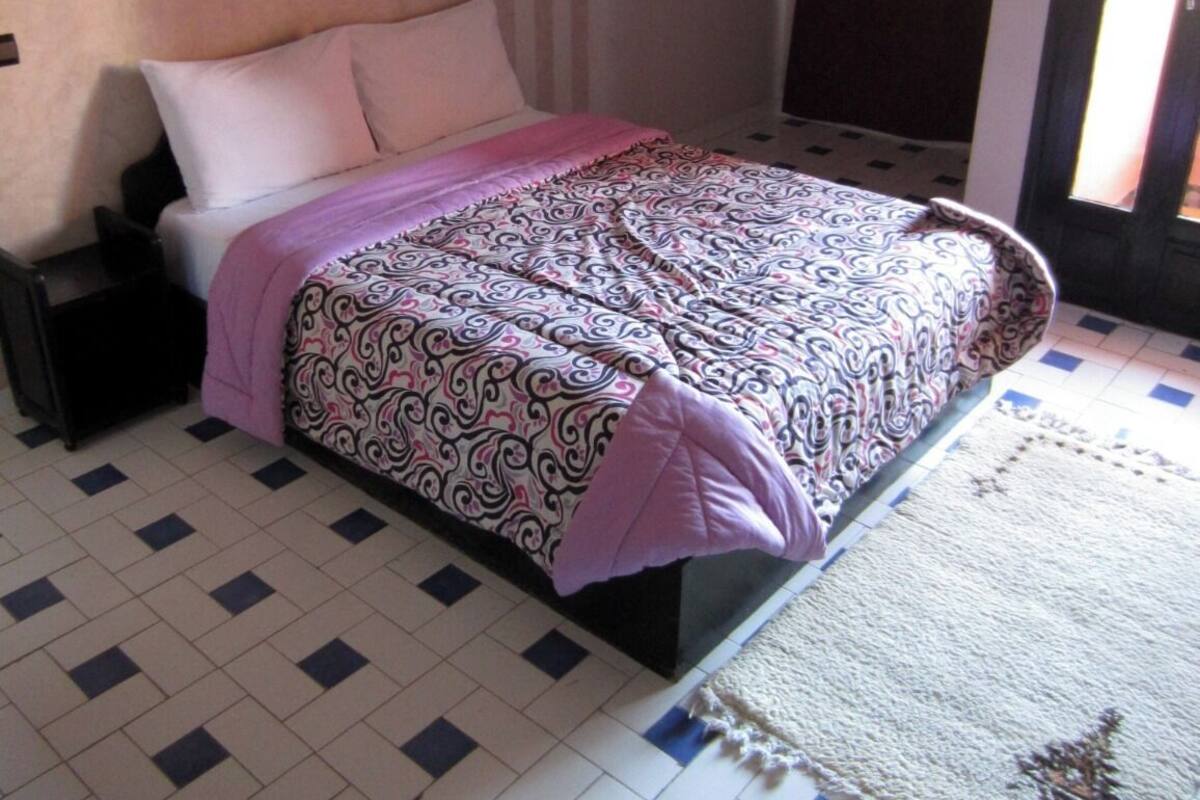 Un dormitorio con cama, mesita de noche, lámpara de pared, ventana con cortinas y alfombra estampada.