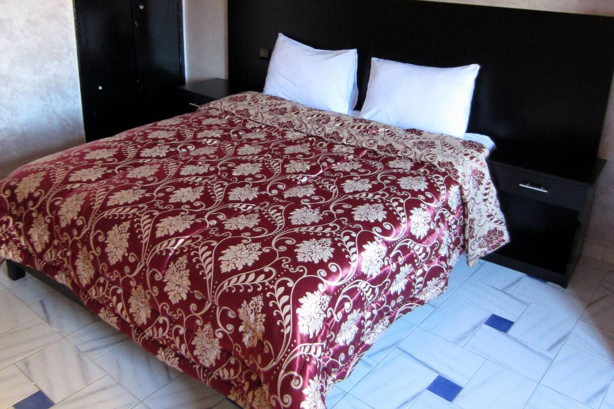 Una cama con colcha estampada, cabecero negro y mesita de noche con lámpara.