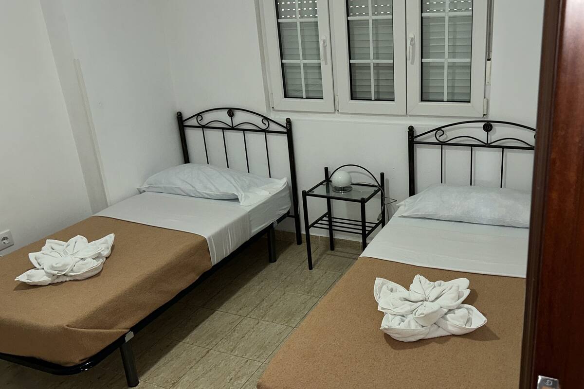 Une petite chambre propre, avec deux lits simples, une petite table et une fenêtre avec des stores.