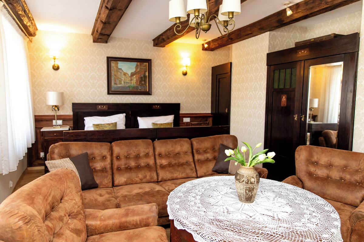 A room with a brown upholstered sofa set, a round table with a lace tablecloth, and a vase with flowers.