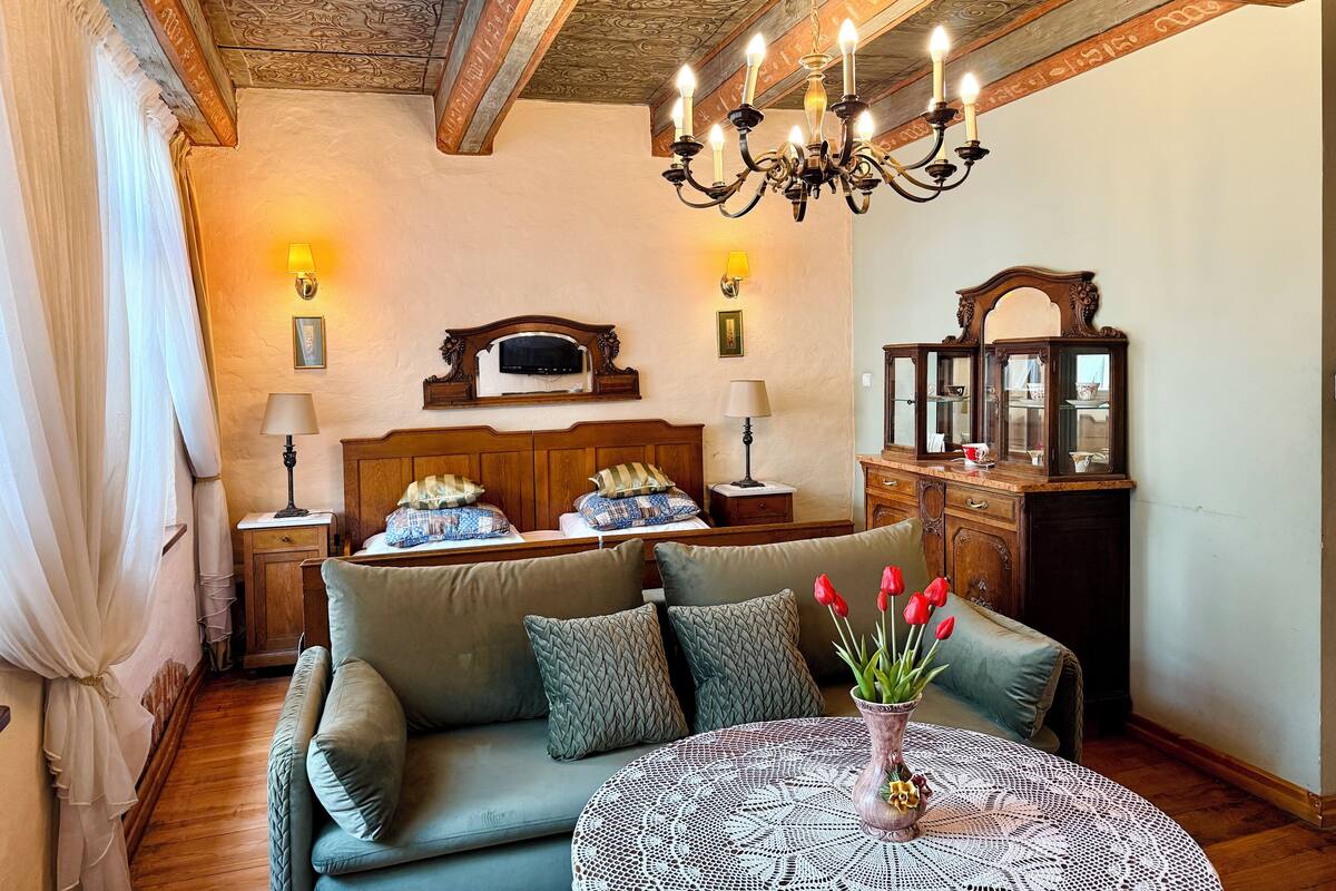 A cozy room with a wooden ceiling, a chandelier, a bed with a wooden headboard, a sofa, a round table with a vase of flowers, and a wooden cabinet with glass doors.