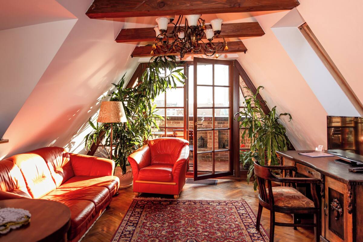 A cozy loft bedroom with a sloped ceiling, a red leather sofa, a wooden desk, and a large window with a view.