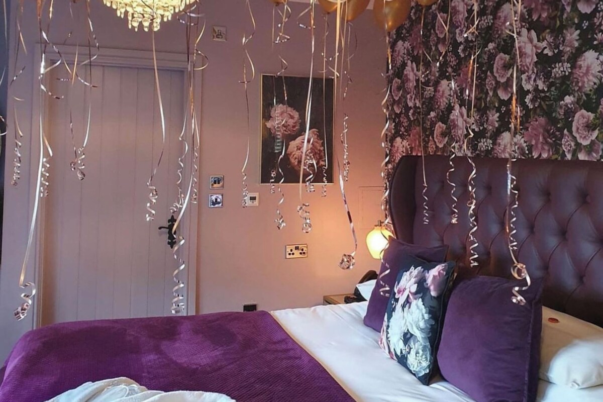 A bedroom with a bed, purple bedding, a chandelier, and gold balloons.
