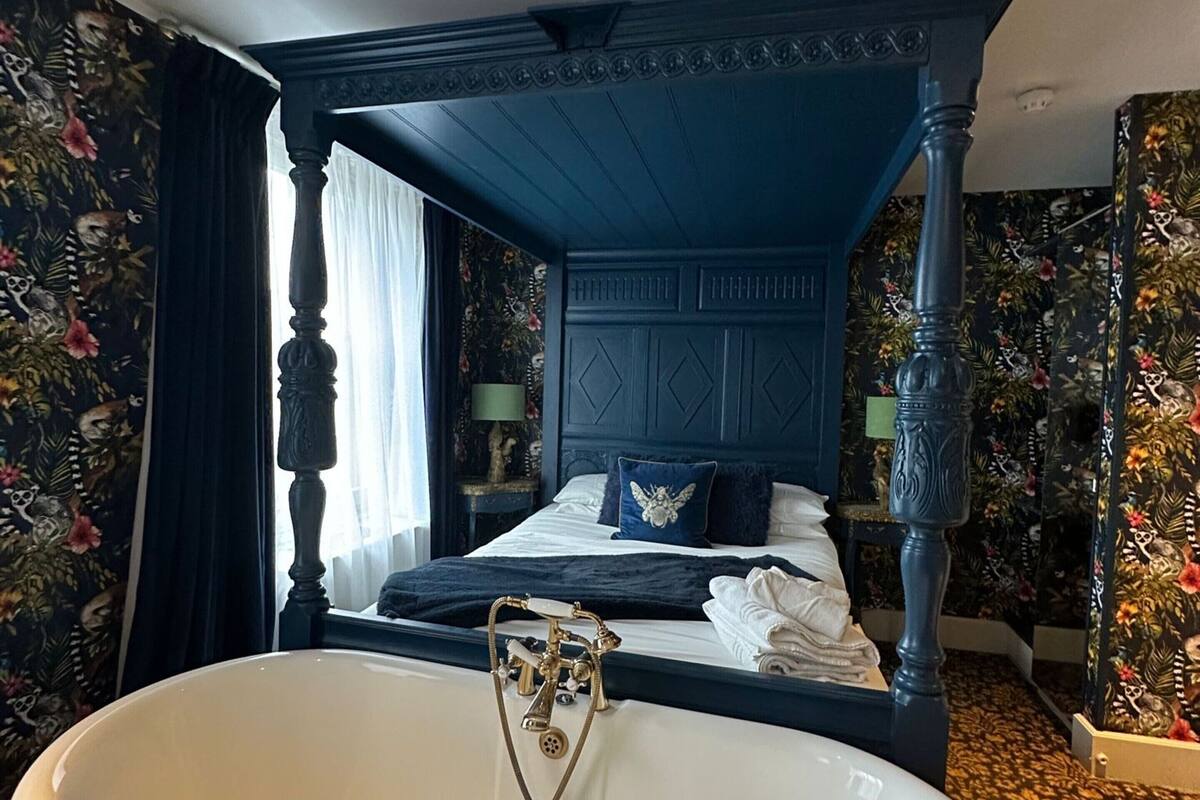 A four-poster bed with a dark blue canopy, a white bathtub with a vintage faucet, and floral wallpaper.