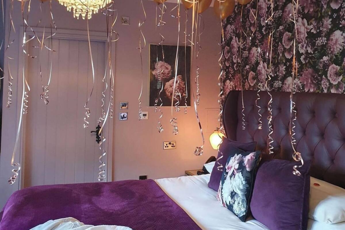 A bedroom with a bed, purple bedding, a chandelier, and gold balloons.