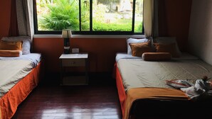 A room with two beds, a window with a view of greenery, a bedside table with a lamp, and a small nightstand. - Hotel Your House (Rio Segundo)