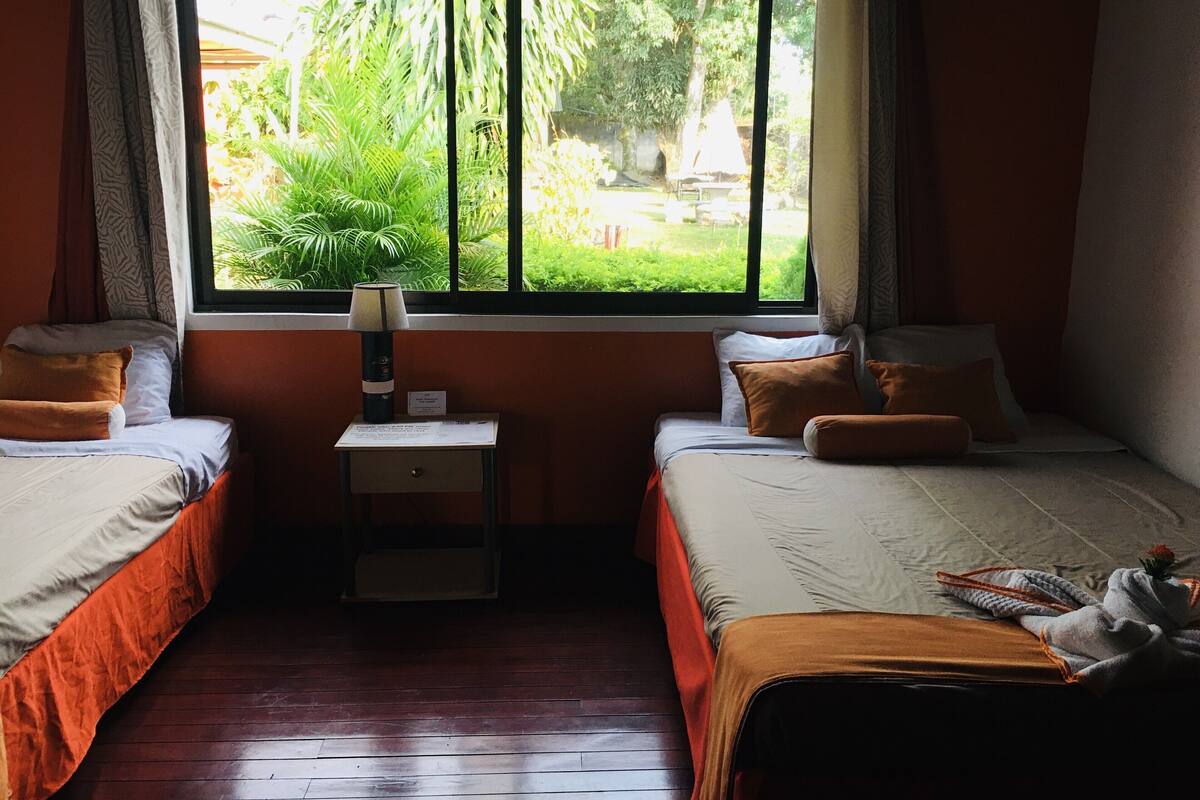 A room with two beds, a window with a view of greenery, a bedside table with a lamp, and a small nightstand.