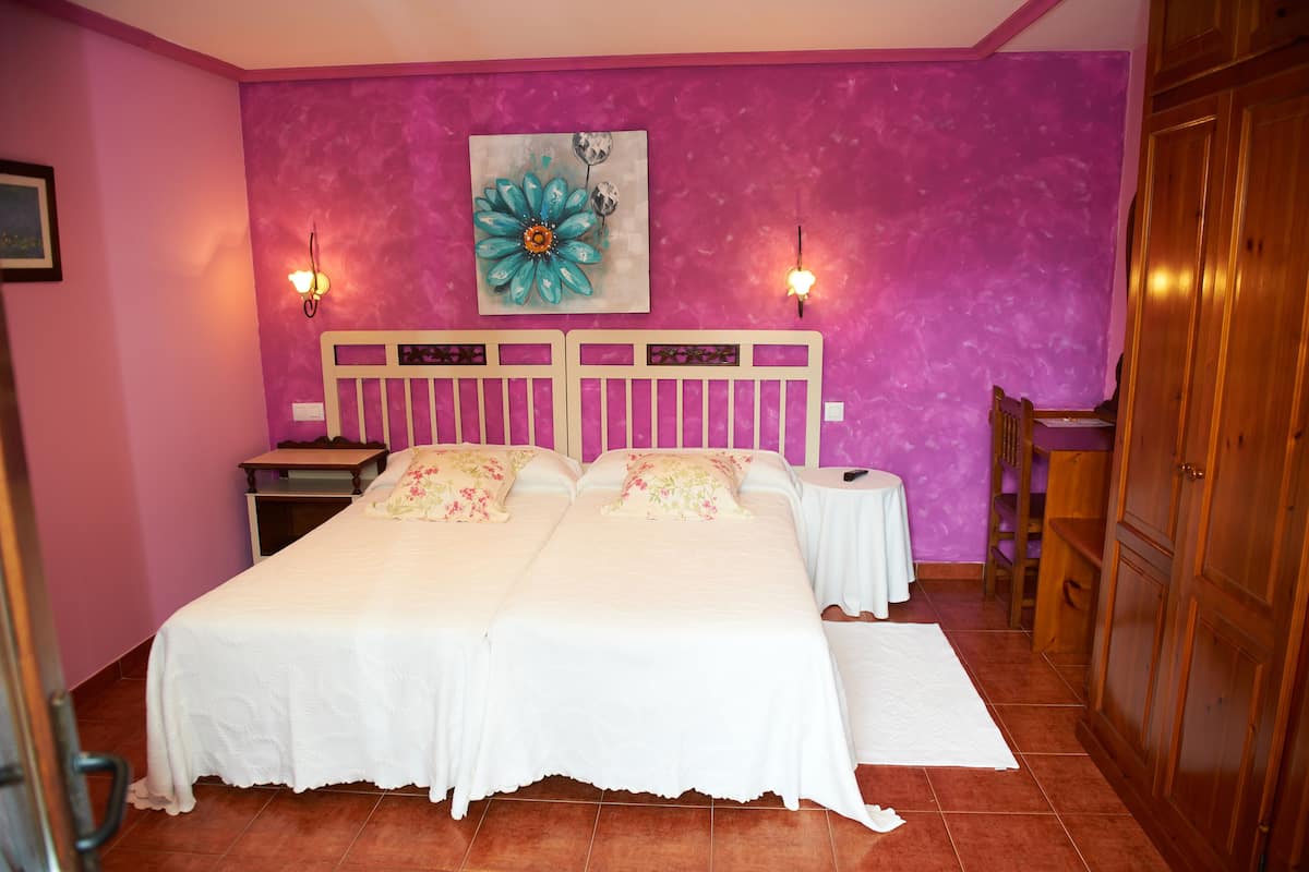A double bed with white linens, a nightstand, a small table, and a wooden wardrobe in a room with pink walls and a large floral painting.