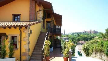 Un edificio amarillo con una escalera que conduce a un balcón, rodeado de vegetación y vistas a una colina con edificios.