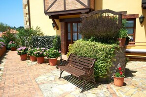 A stone-paved patio with a wooden bench, surrounded by potted plants and a yellow building with a brick facade.