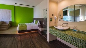 Un dormitorio moderno con cama, pared de acento verde, escalera de madera y baño con espejo grande y mosaicos verdes.