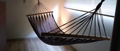 A hammock suspended from the ceiling in a room with wooden flooring and a window with blinds.