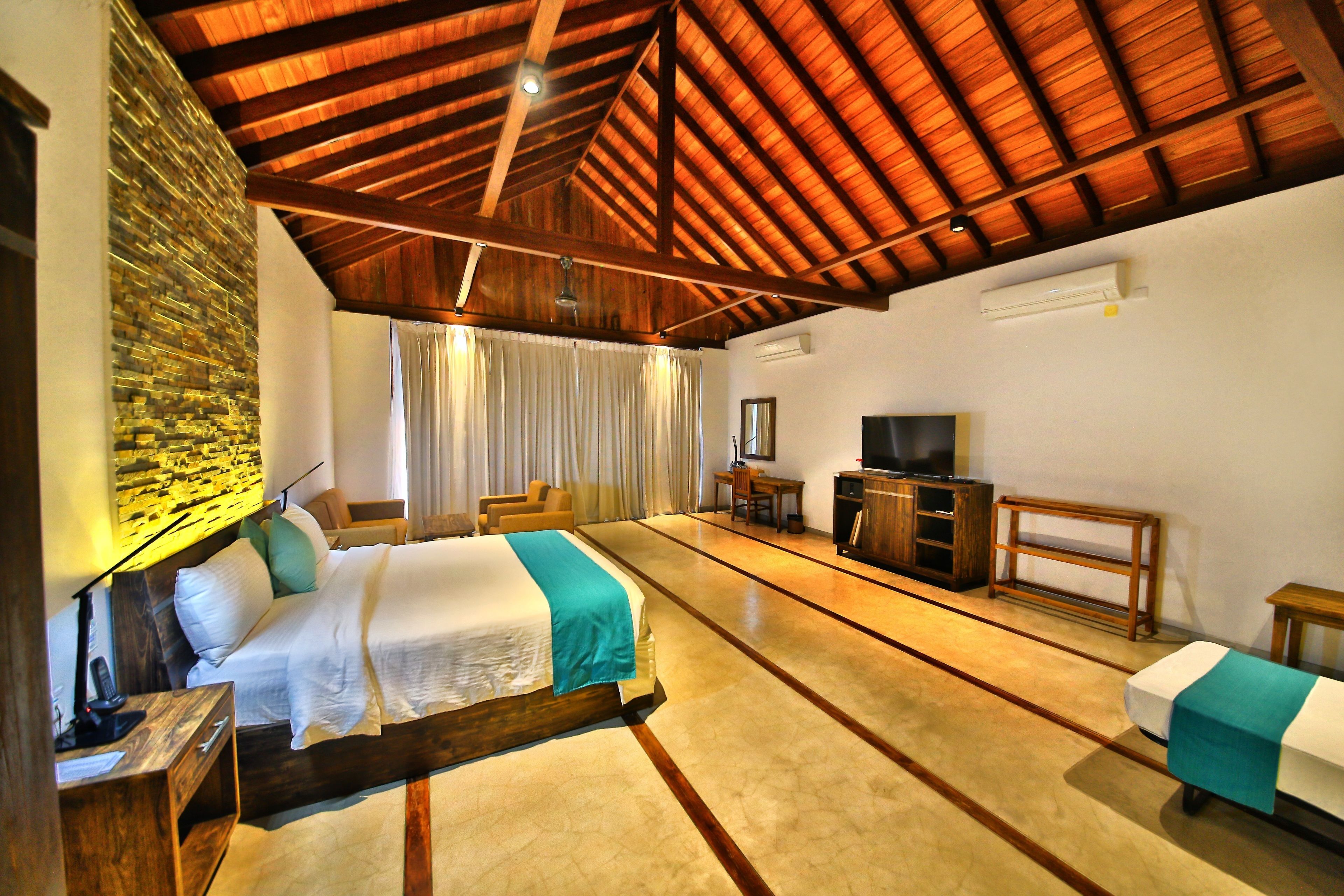 Amplia habitación de hotel con una cama grande, techo de madera, pared de piedra, televisor y escritorio.