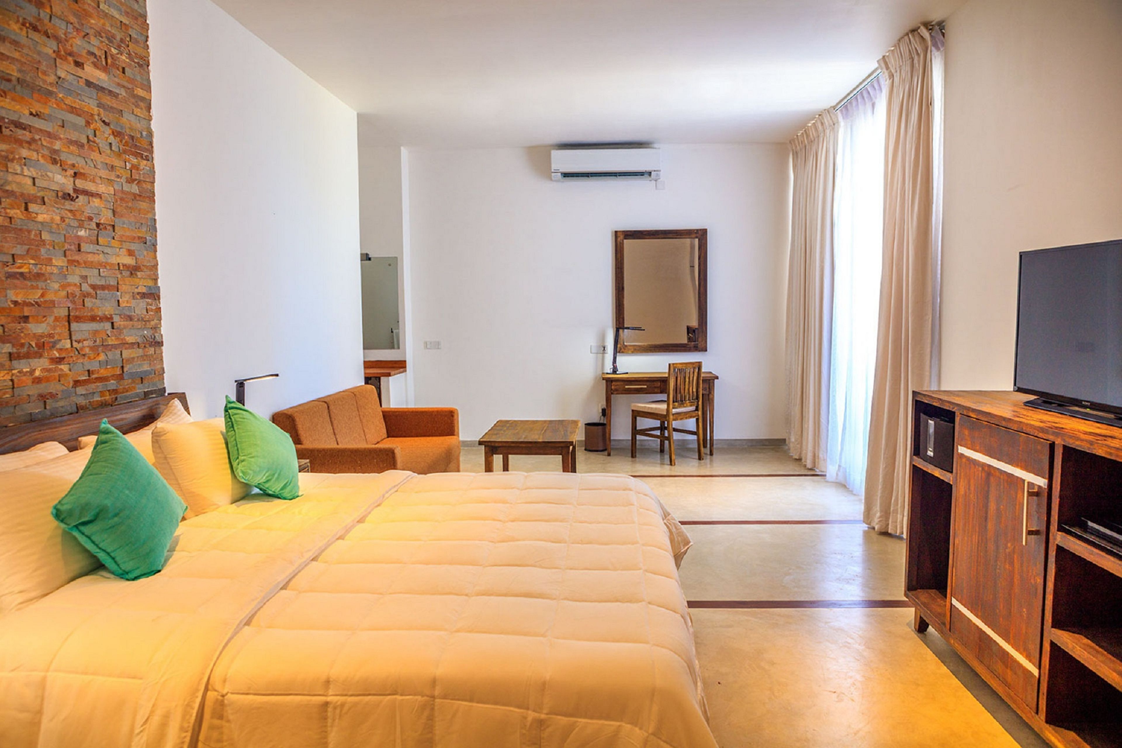 A hotel room with a large bed, a wooden desk, a chair, a television, and a brick wall.