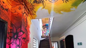 Un escalier orné d’une fresque colorée sur le mur, menant à une pièce où se trouvent un lit et une fenêtre.