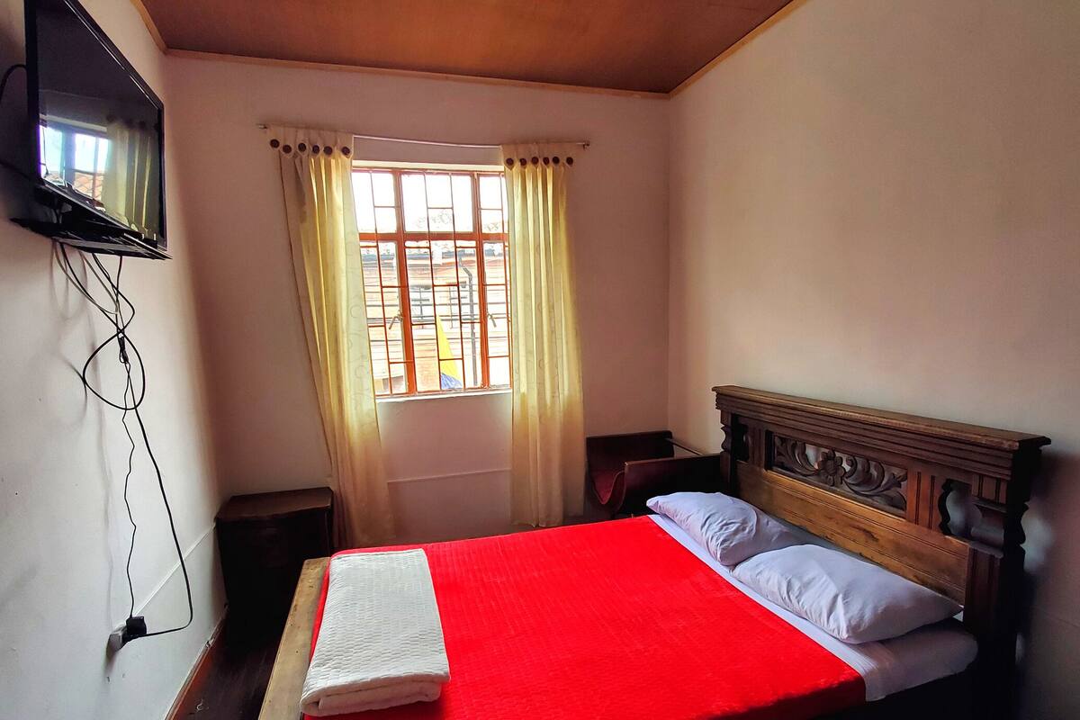 Een slaapkamer met een enkel bed, een aan de muur bevestigde televisie en een raam met gordijnen.