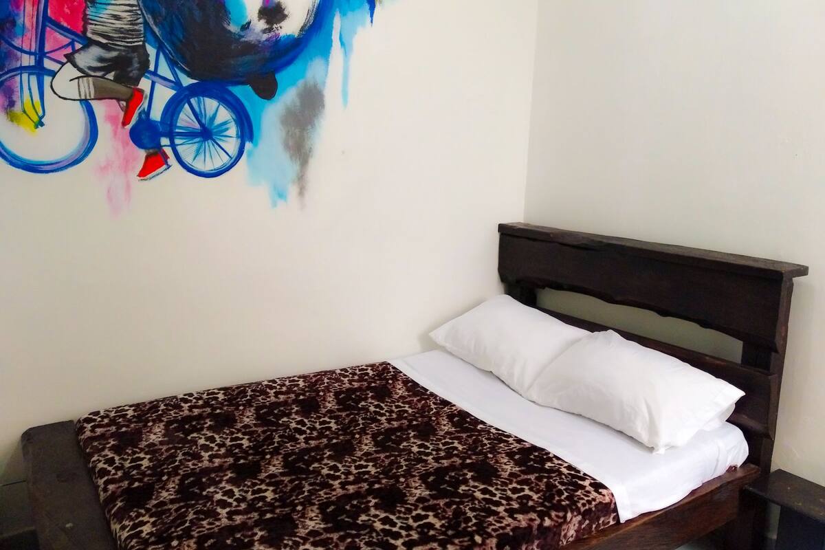 Een bed met een luipaardprint deken, witte kussens, een donker houten hoofdbord en een kleurrijke abstracte muurschildering.