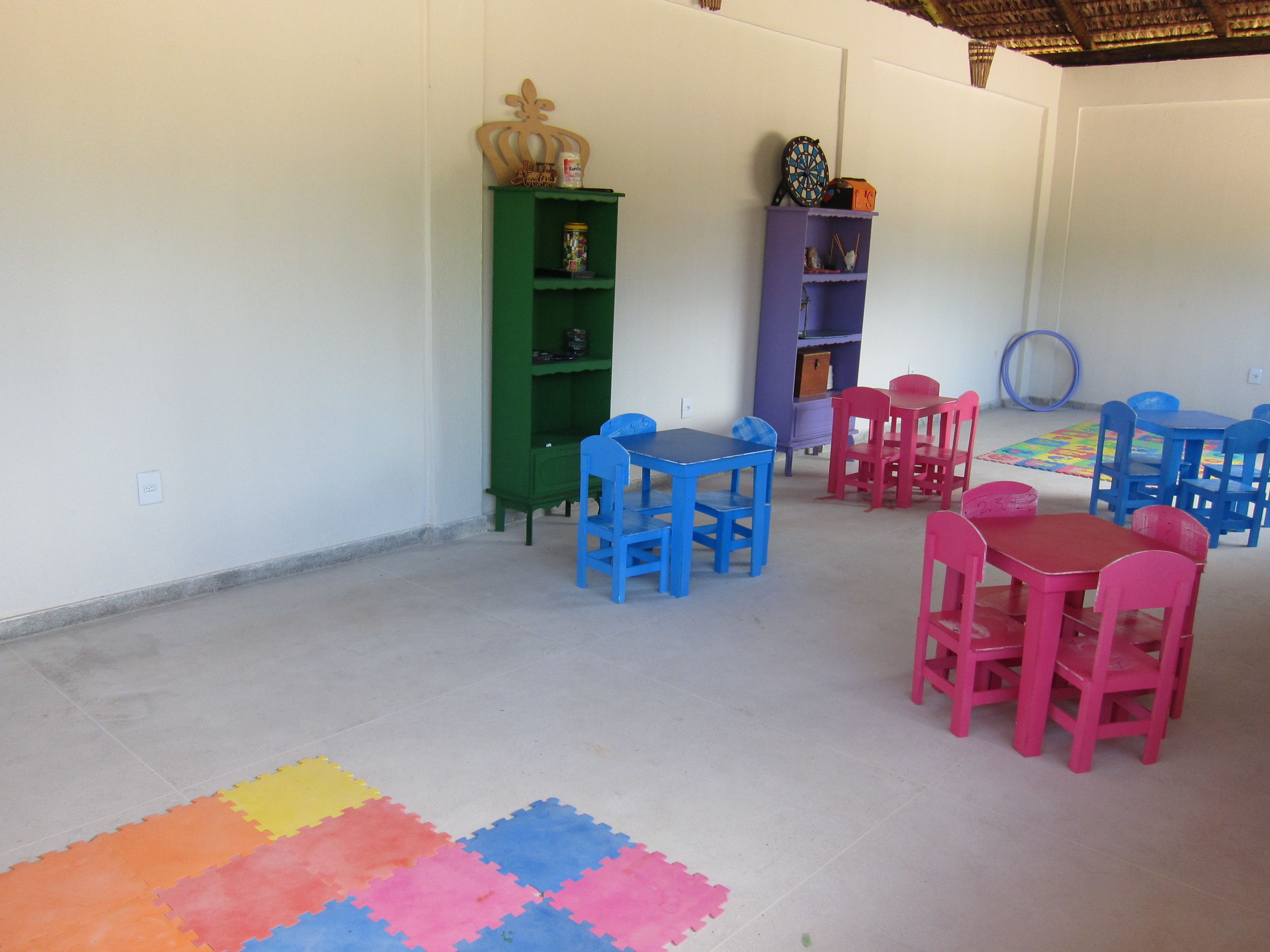 Una habitación con suelo de baldosas, un tapete de juegos de colores y una mesa con sillas. Hay dos estanterías con varios objetos encima.