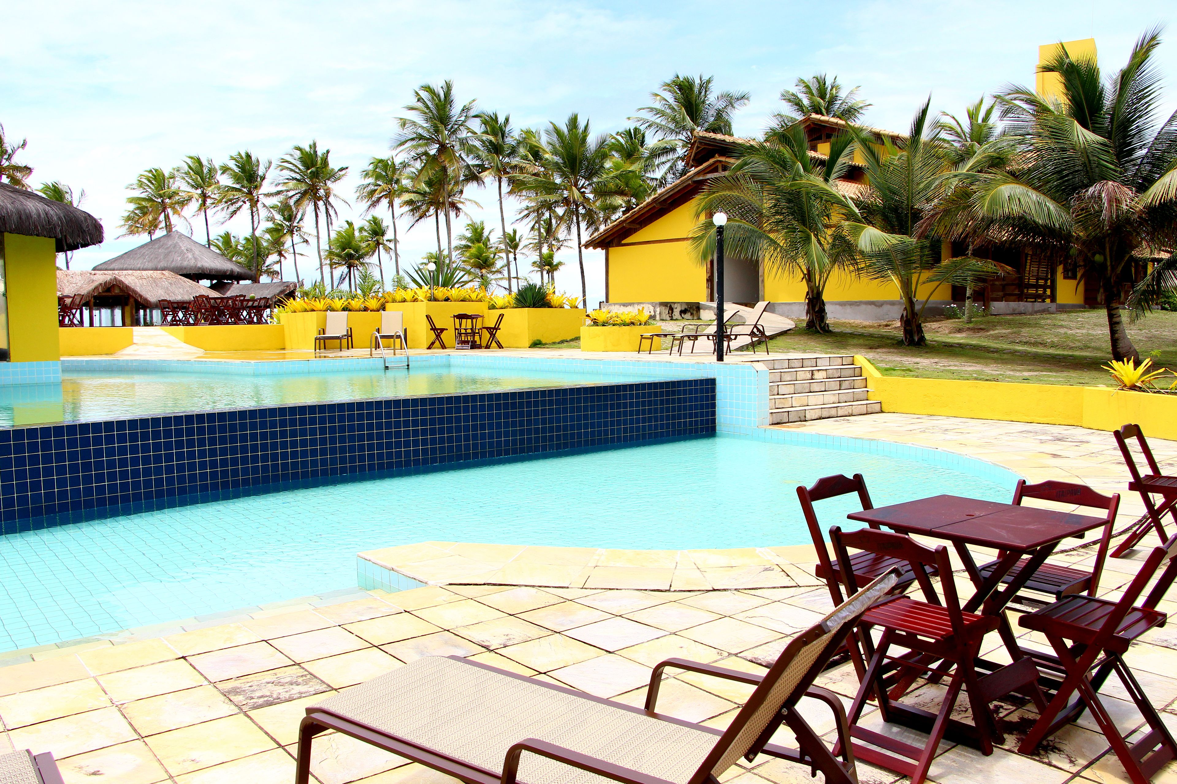 A pool area with tiled flooring, wooden lounge chairs, and a yellow building with a thatched roof.