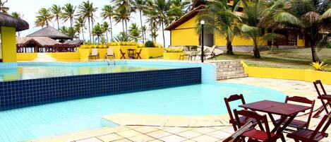 A pool area with tiled flooring, wooden lounge chairs, and a yellow building with a thatched roof.