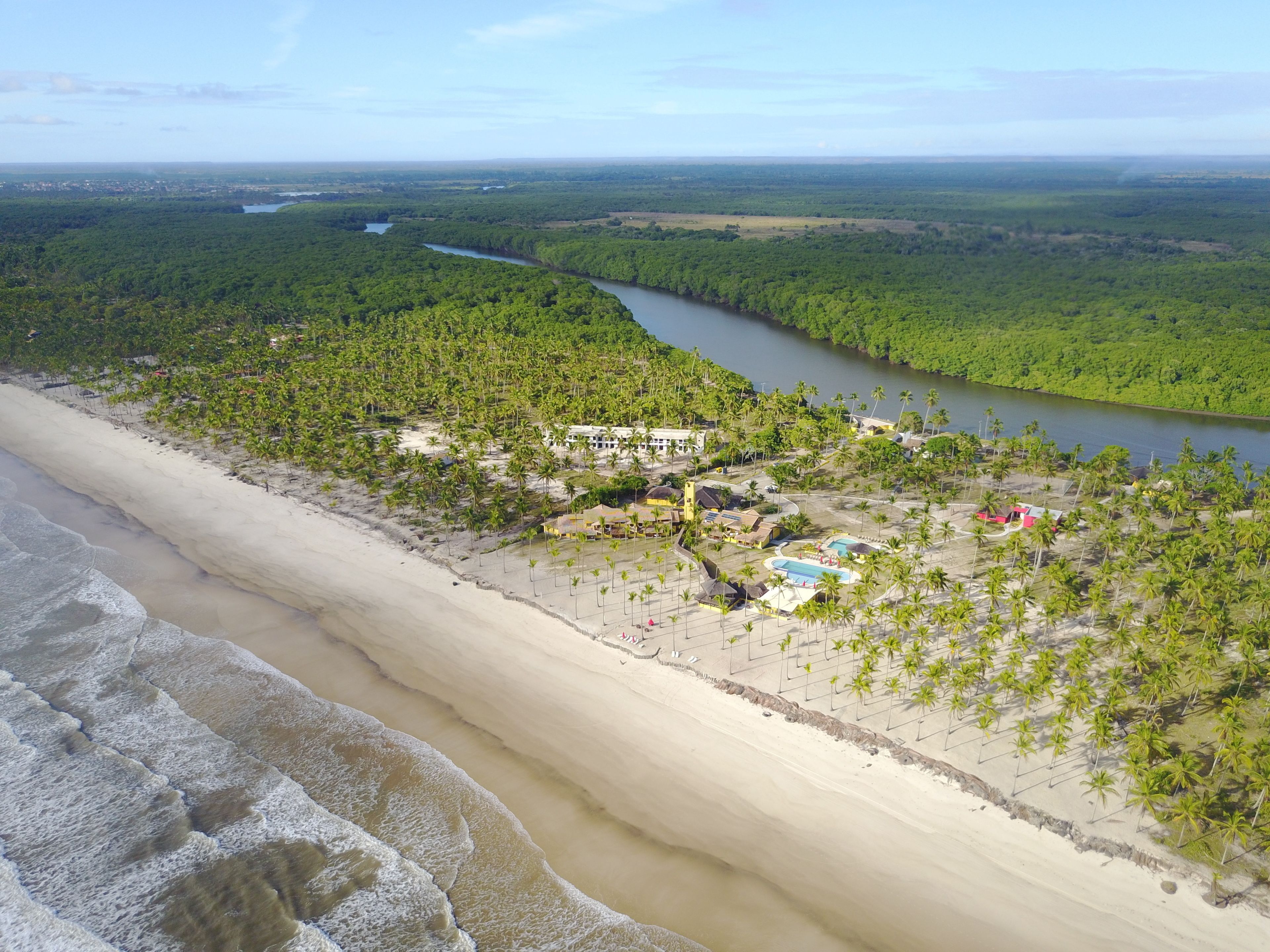 Vista aérea de un complejo turístico frente al mar, con playa, palmeras y un río.