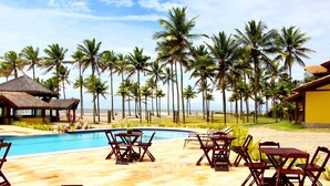 Un área junto a la piscina con sillones de descanso y una estructura de techo de paja, rodeada de palmeras y una vista despejada de la playa.