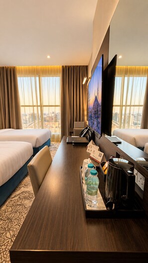A hotel room with two beds, a desk with a laptop, a television, and a cityscape view.