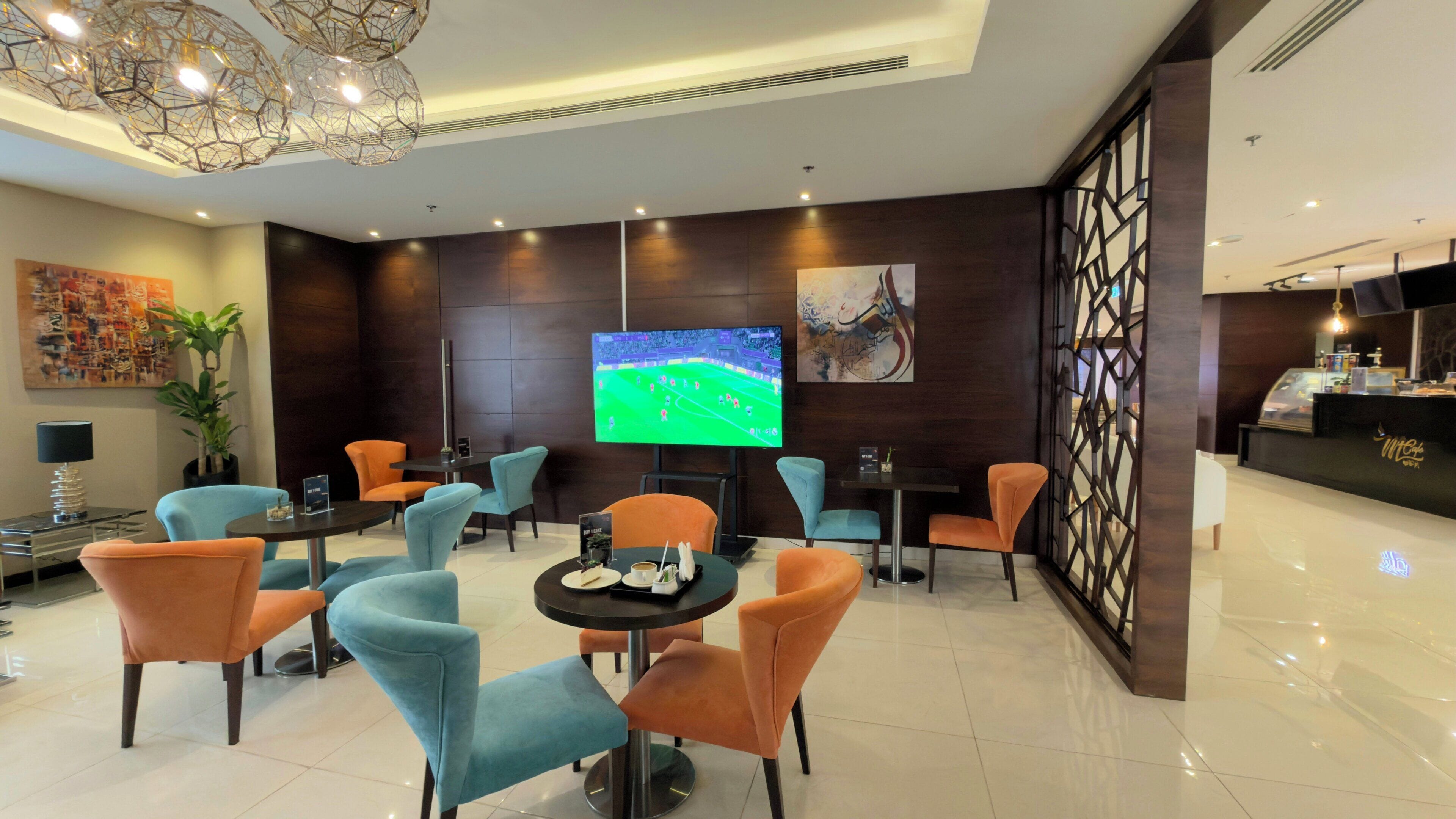 A lobby with a TV displaying a sports game, seating areas with orange and blue chairs, a reception desk, and a decorative wall.