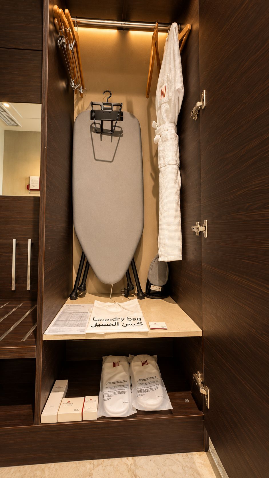 A hotel room with a wooden wardrobe, a hanging rack with towels, and a shelf with a laundry bag and toiletries.