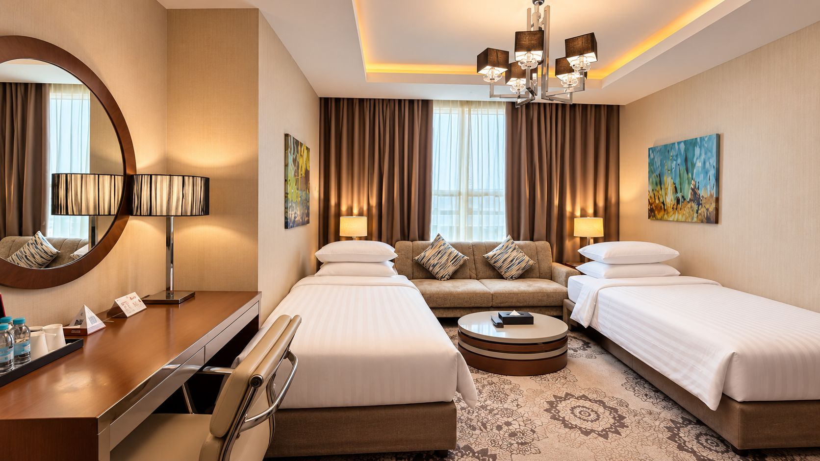 A hotel room with two beds, a sofa, a desk, a chair, a round coffee table, and a large mirror.