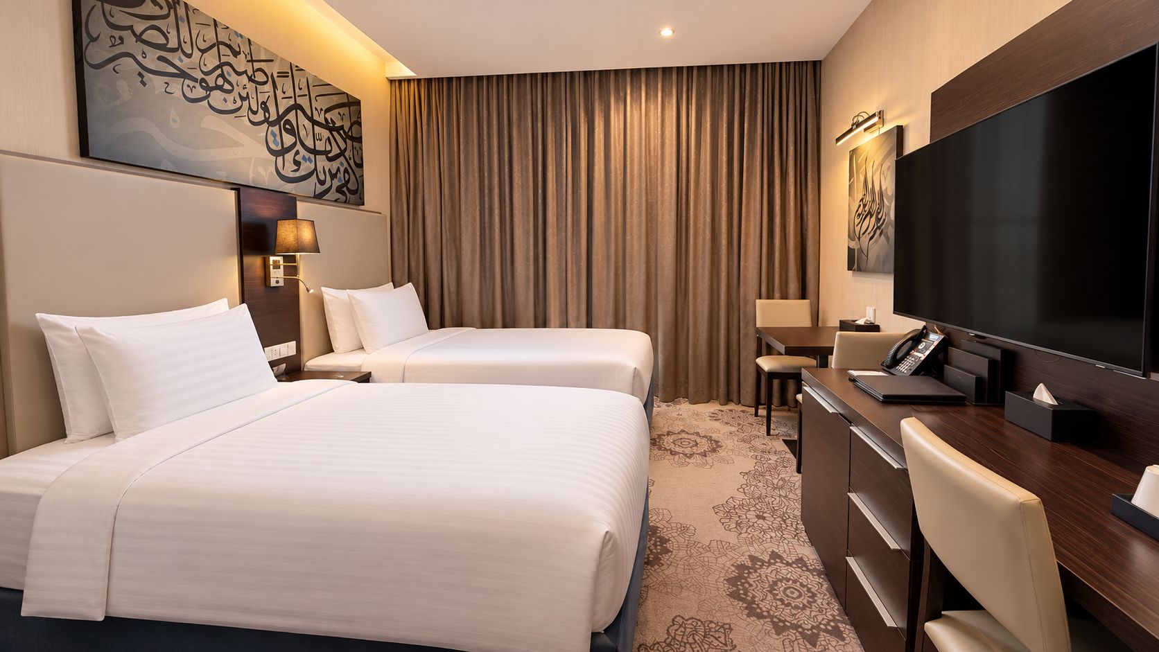 A hotel room with two beds, a desk with a telephone, a flat-screen TV, and artwork on the walls.