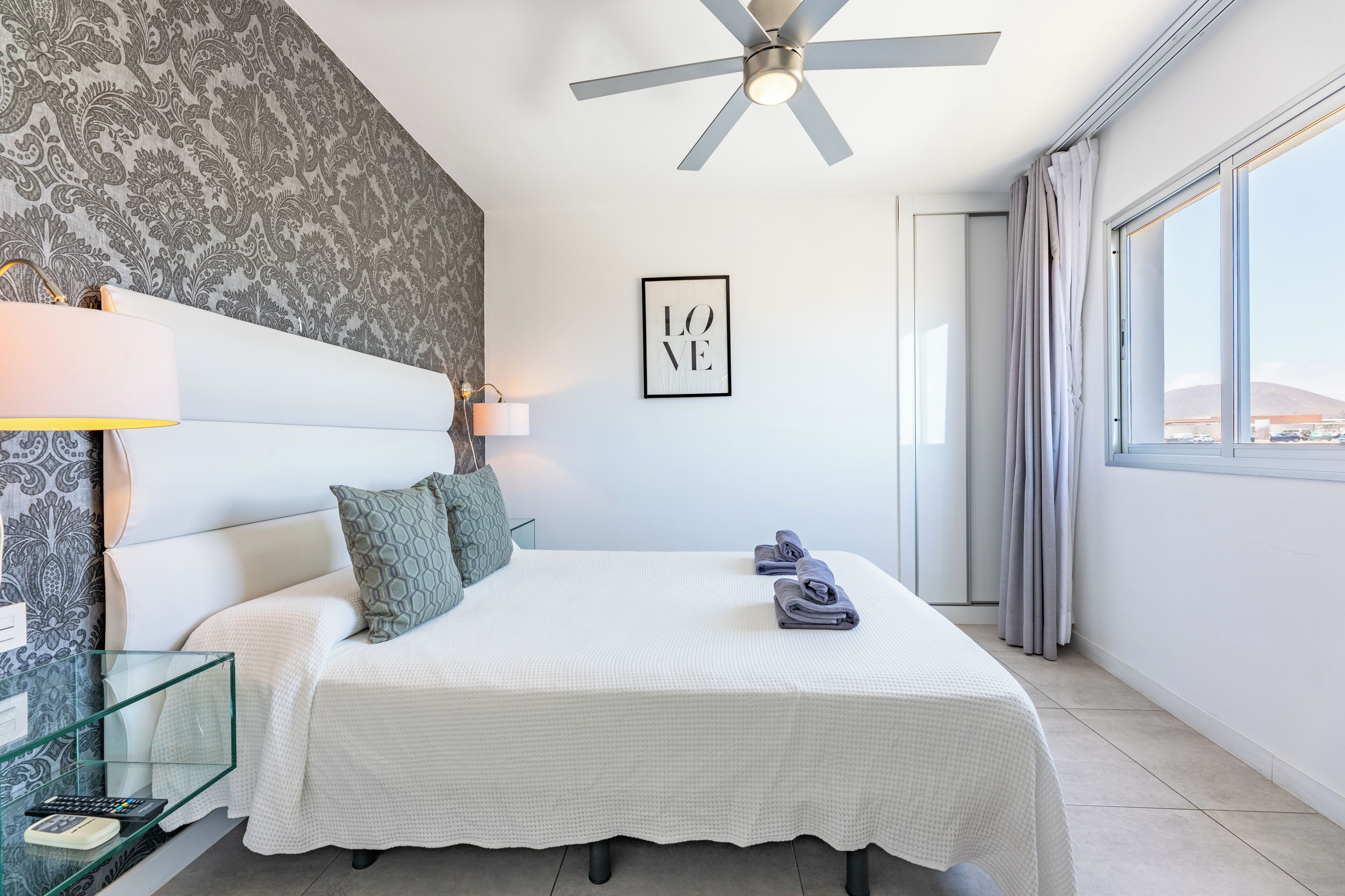 A hotel room with a bed, a ceiling fan, a framed 'LOVE' sign, and a view of the outside.