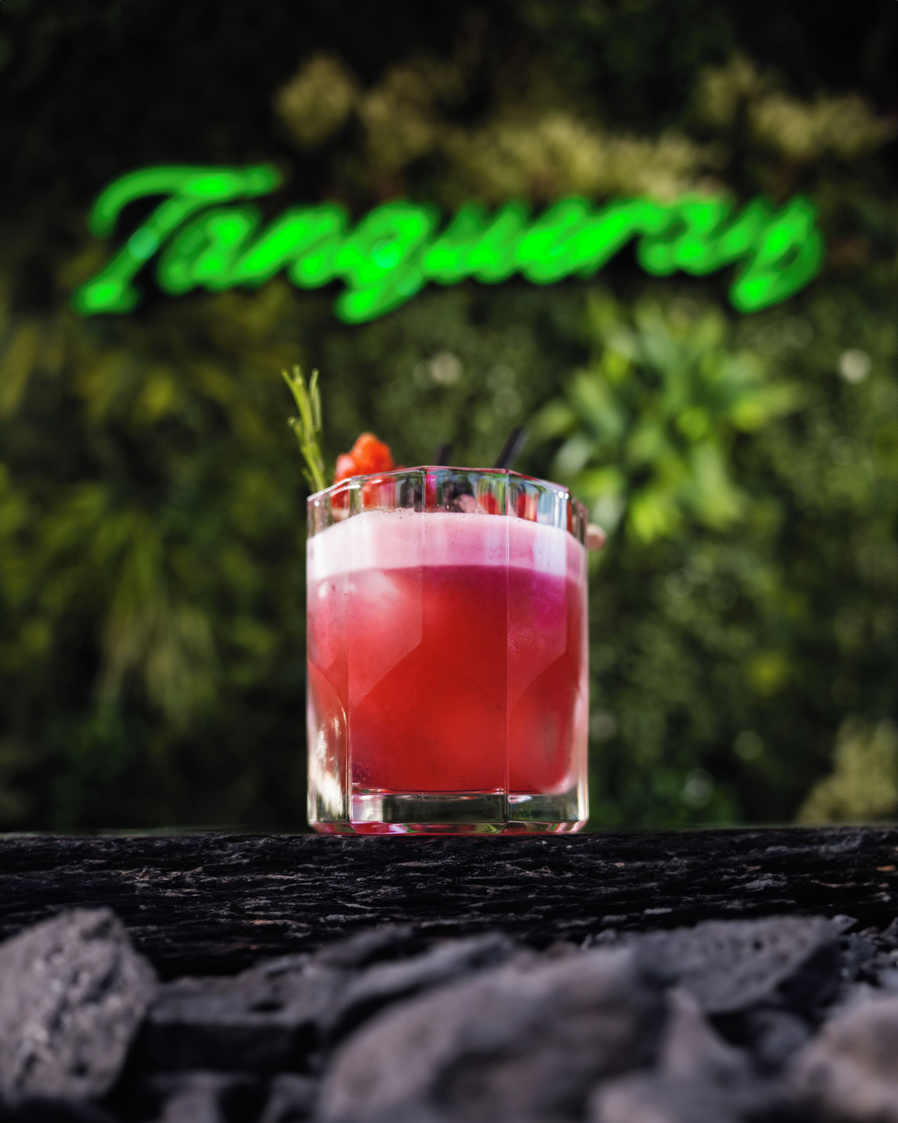 A cocktail glass with a layered drink, garnished with a sprig and a cherry, on a wooden surface.
