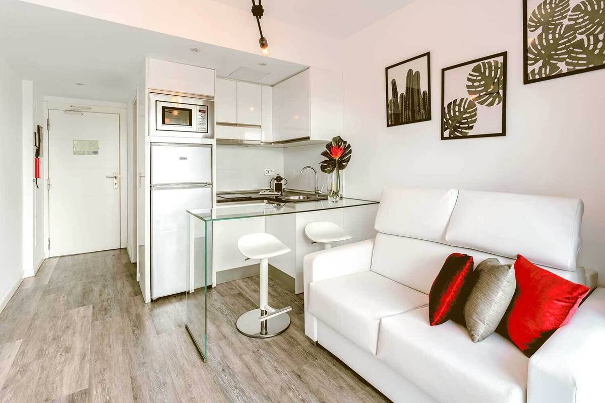 Una cocina moderna con armarios blancos y una isla de vidrio, un sofá blanco con cojines rojos y grises, y obras de arte enmarcadas en la pared.