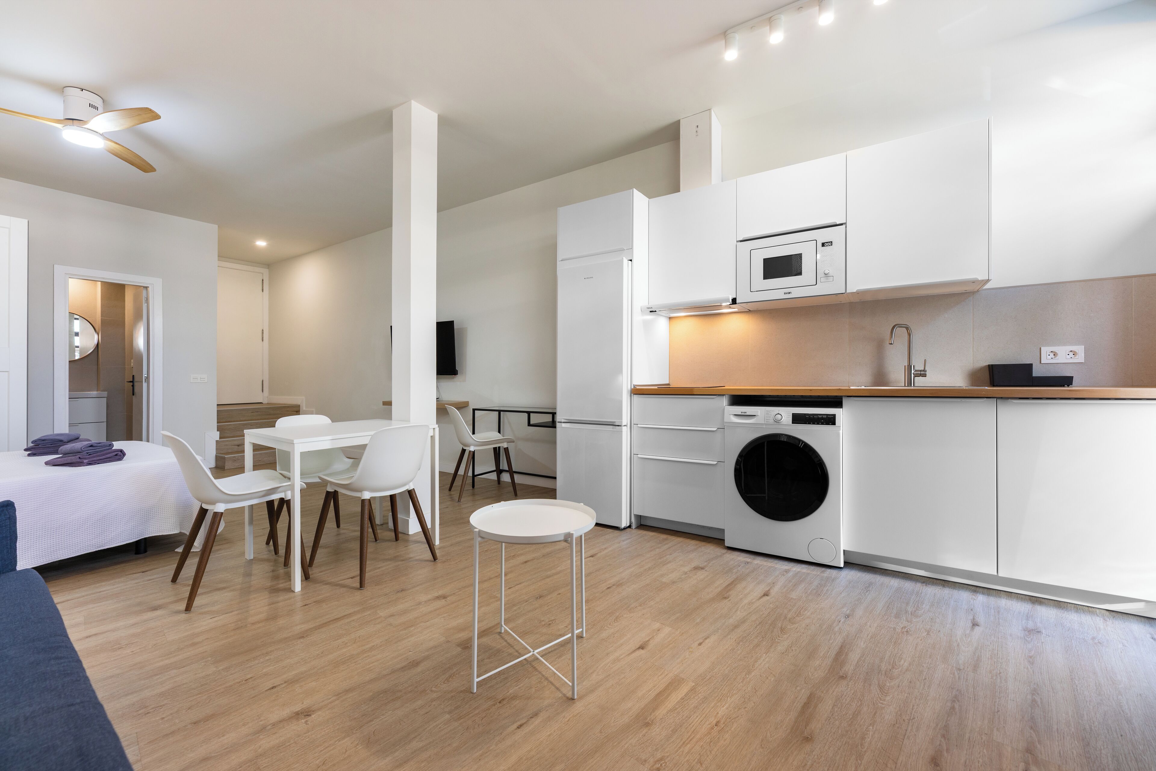 Cocina moderna con armarios blancos, microondas y lavadora. Cuenta con un comedor con mesa y sillas.