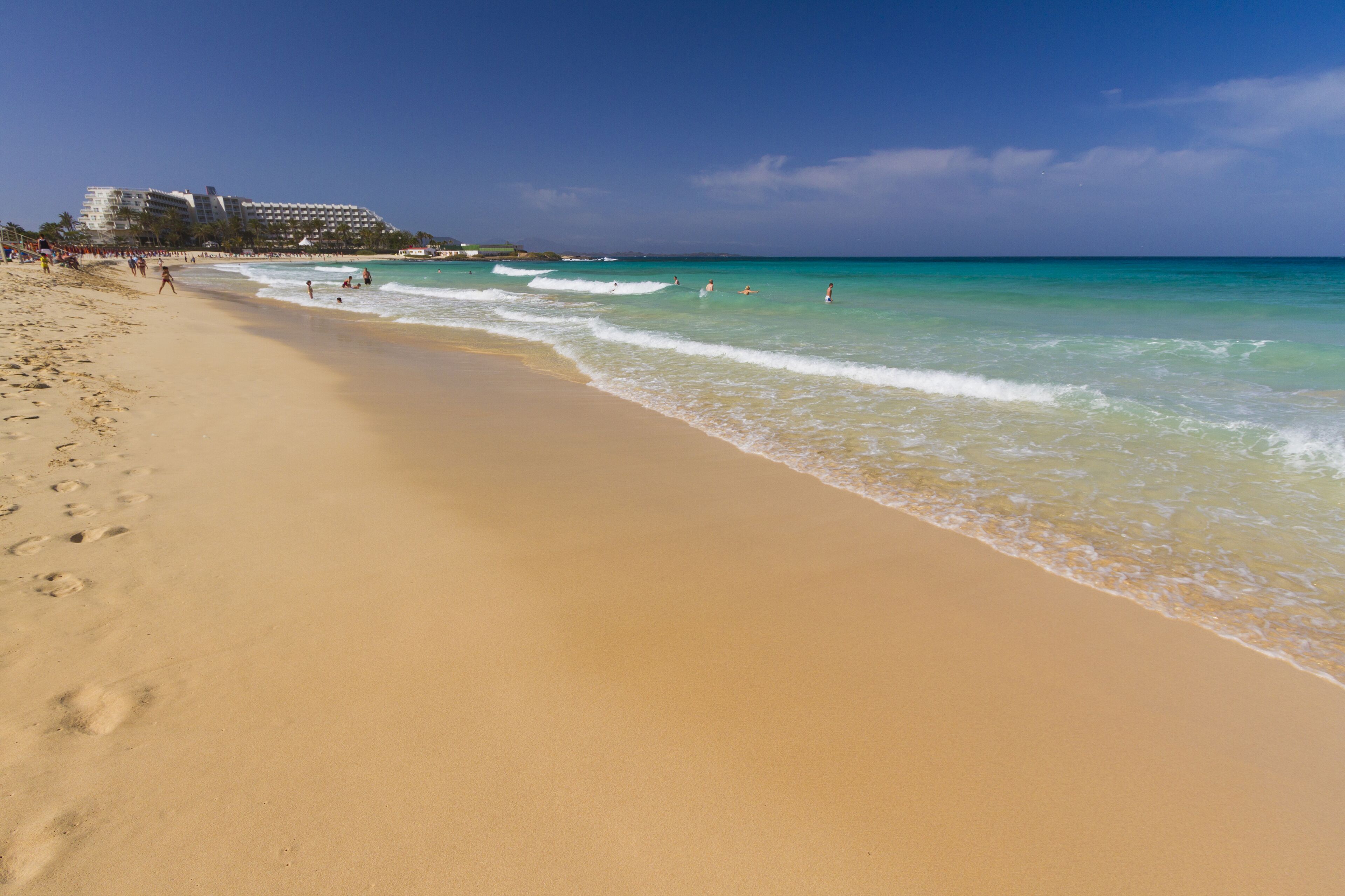 A wide sandy beach with gentle waves, a clear blue sky, and a resort building in the background.