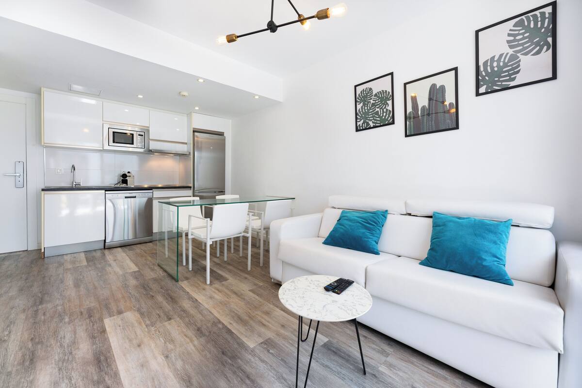 Una sala de estar moderna con un sofá blanco, una mesa de comedor de vidrio y una cocina al fondo.
