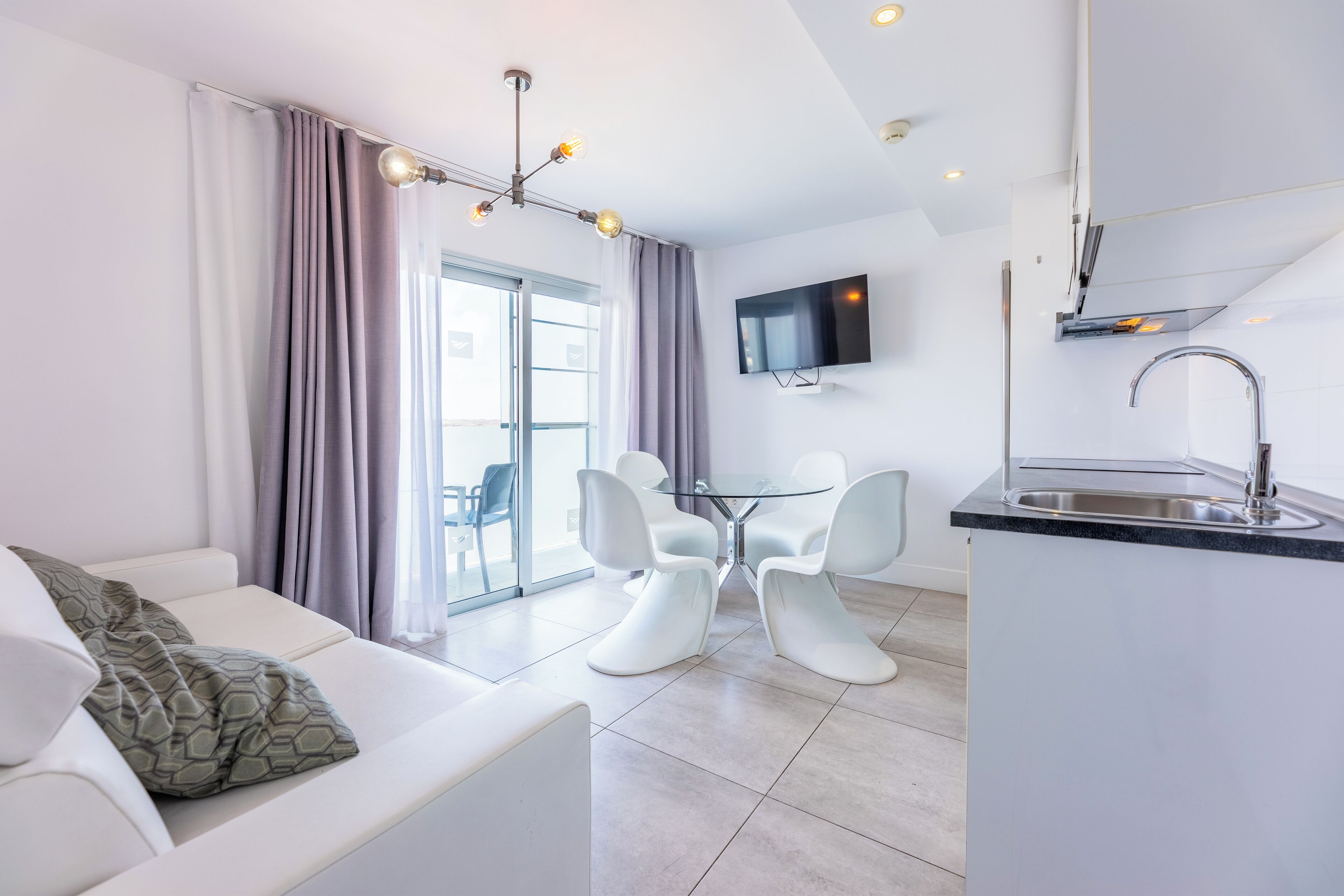 A modern living room with a sofa, a dining table with chairs, a kitchen area with a sink, and a TV mounted on the wall.