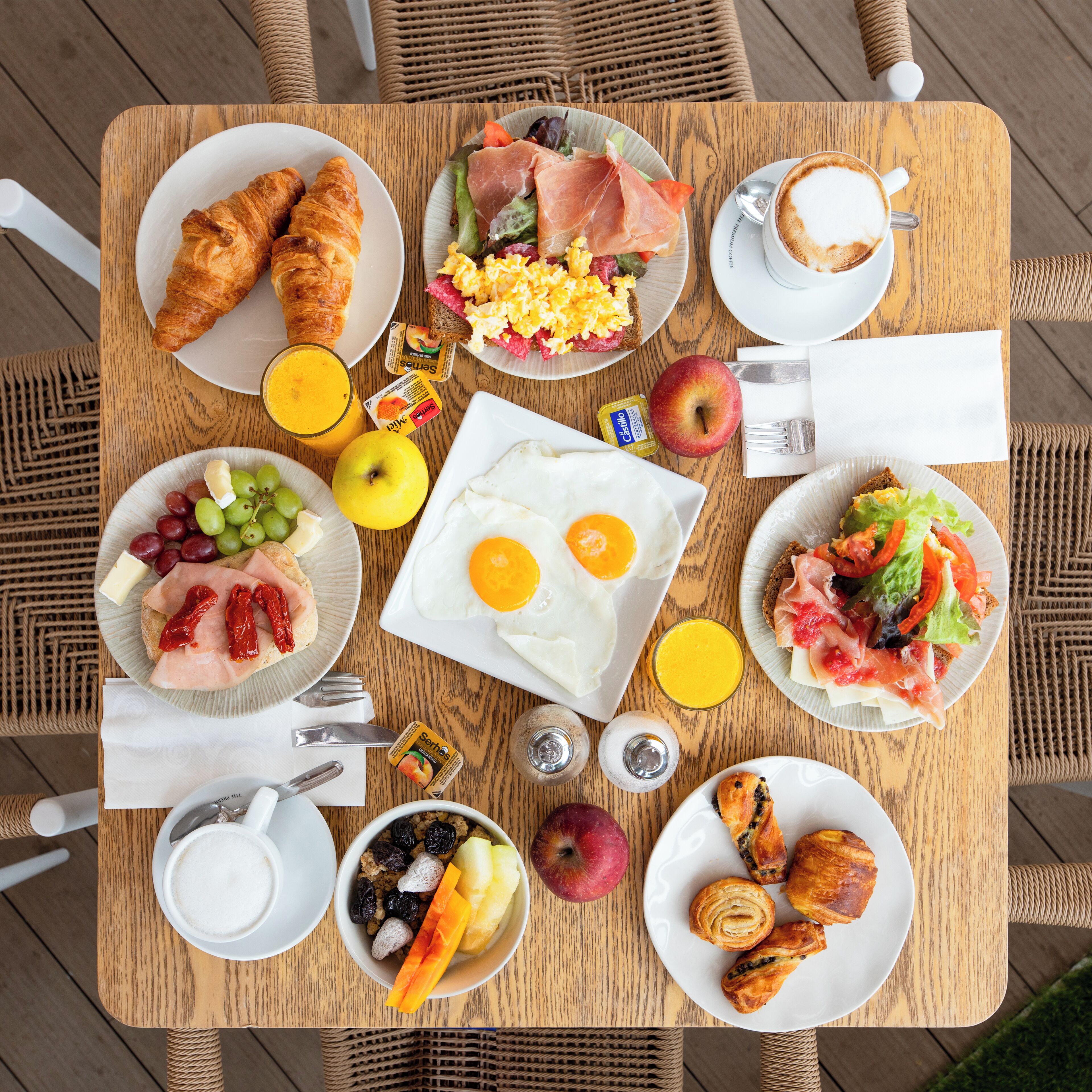 Una mesa de desayuno con cruasanes, huevos, jamón, queso, frutas y café.