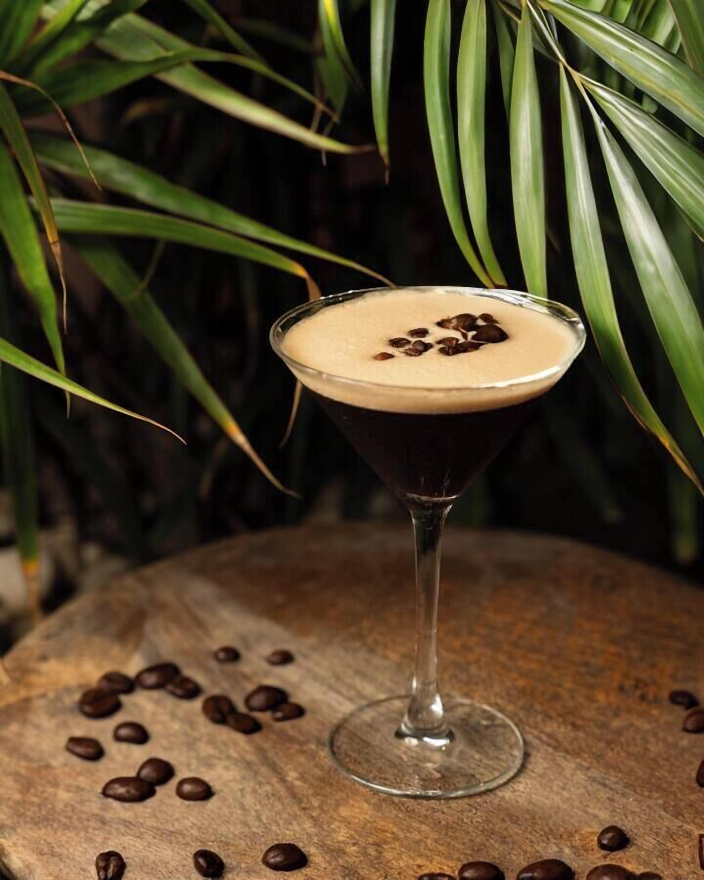 A martini glass with a frothy top, placed on a wooden surface surrounded by coffee beans.