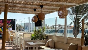 Un espacio de estar al aire libre con mesas y bancos de madera, techo de paja y grandes ventanales que ofrecen vistas de la ciudad.