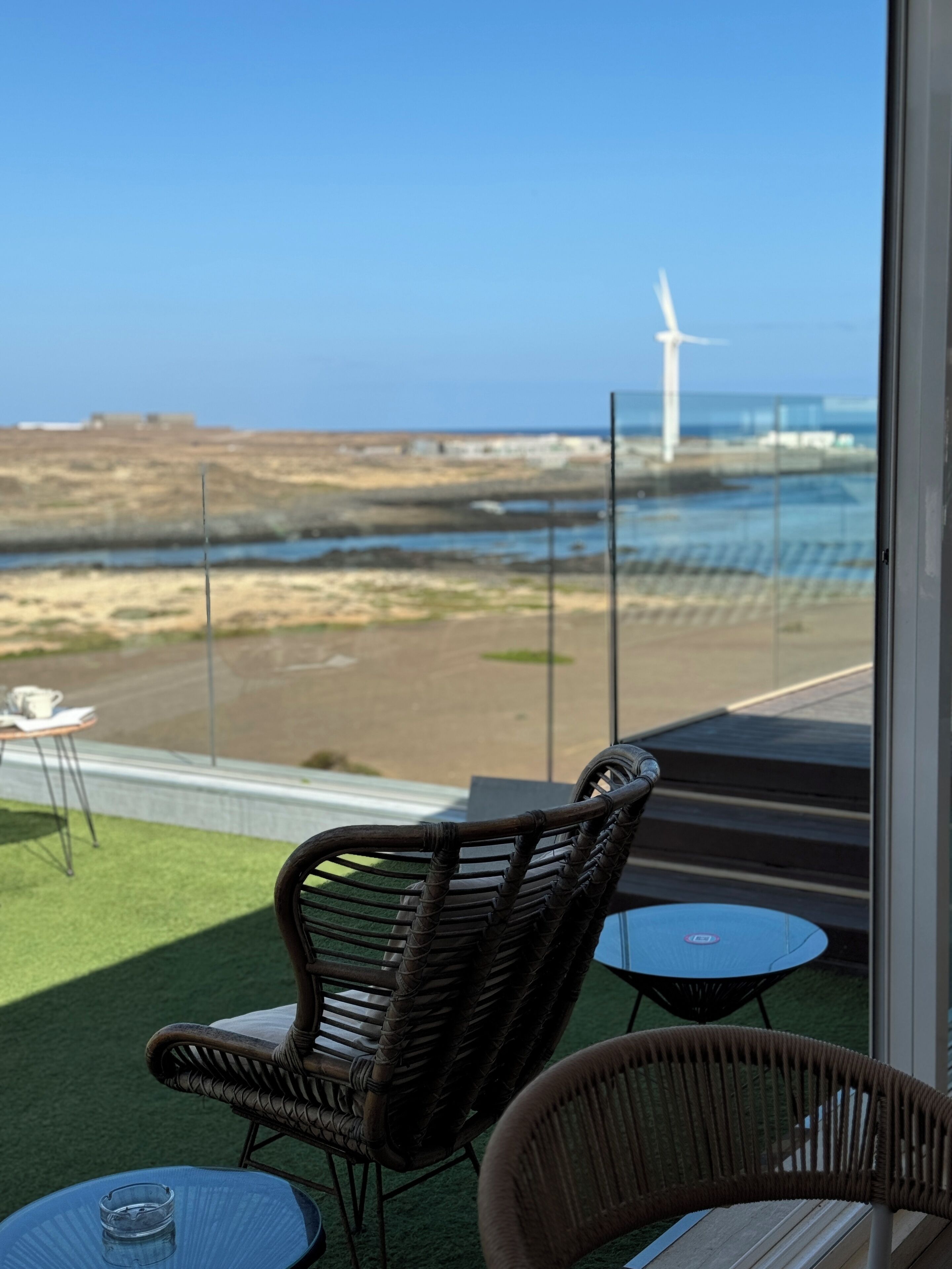 A balcony with wicker chairs and a small table, overlooking a beach with a wind turbine in the distance.